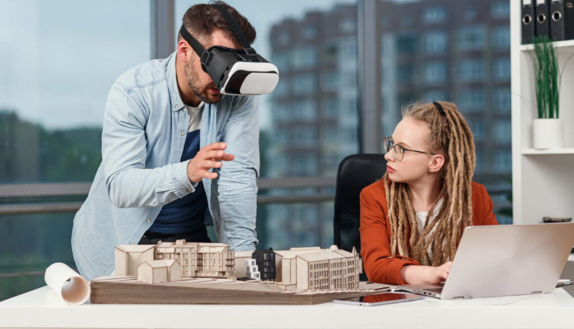 Professional male architect in augmented reality goggles working with mock up of building and woman coworker with laptop