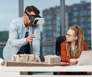 Professional male architect in augmented reality goggles working with mock up of building and woman coworker with laptop