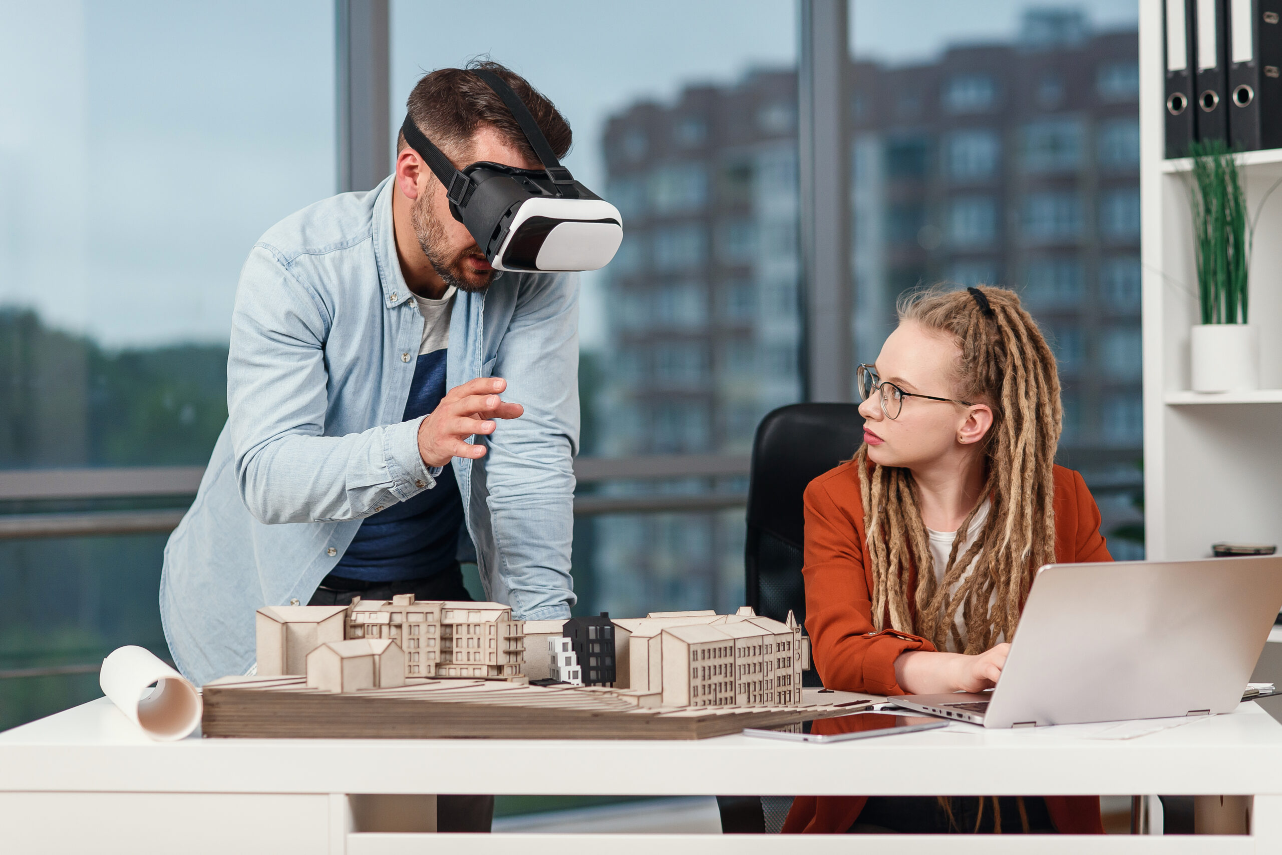 Professional male architect in augmented reality goggles working with mock up of building and woman coworker with laptop